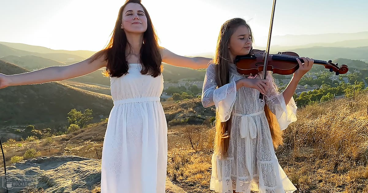 Two young adult females in a scenic outdoor setting with rolling hills and mountains, wearing white dresses; one in a sleeveless dress, the other in a lace dress under warm golden light.