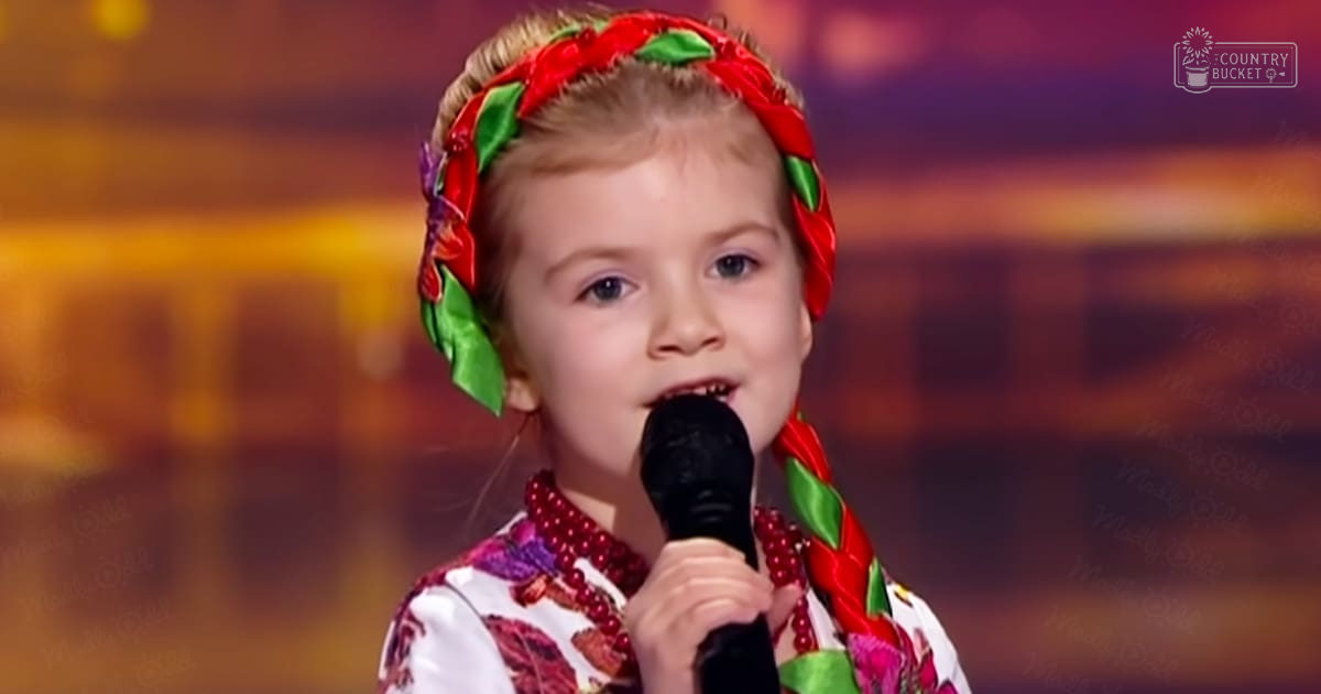A young female child performs on stage in a talent show setting. She wears a white patterned outfit with flowers and leaves, accessorized with a red and green braided headband and a beaded necklace.