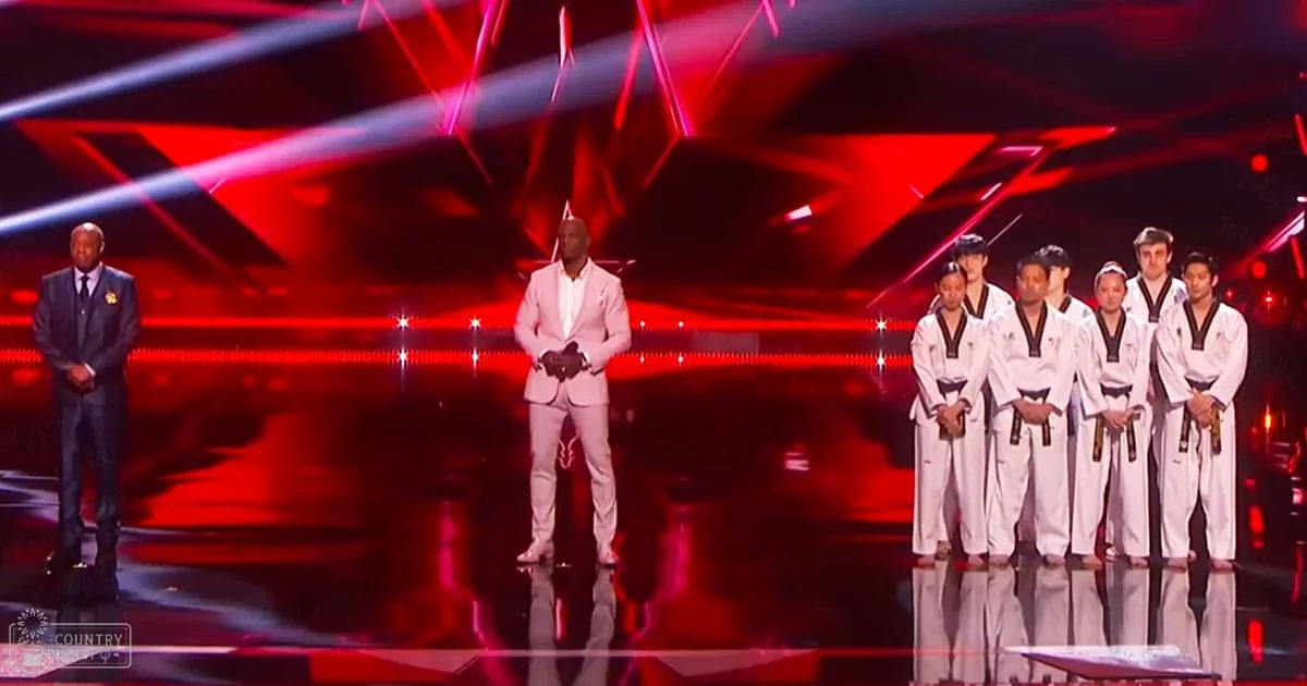 Stage with reflective flooring, red lighting effects, and a person in a light-colored suit standing center stage at an awards ceremony or performance event.