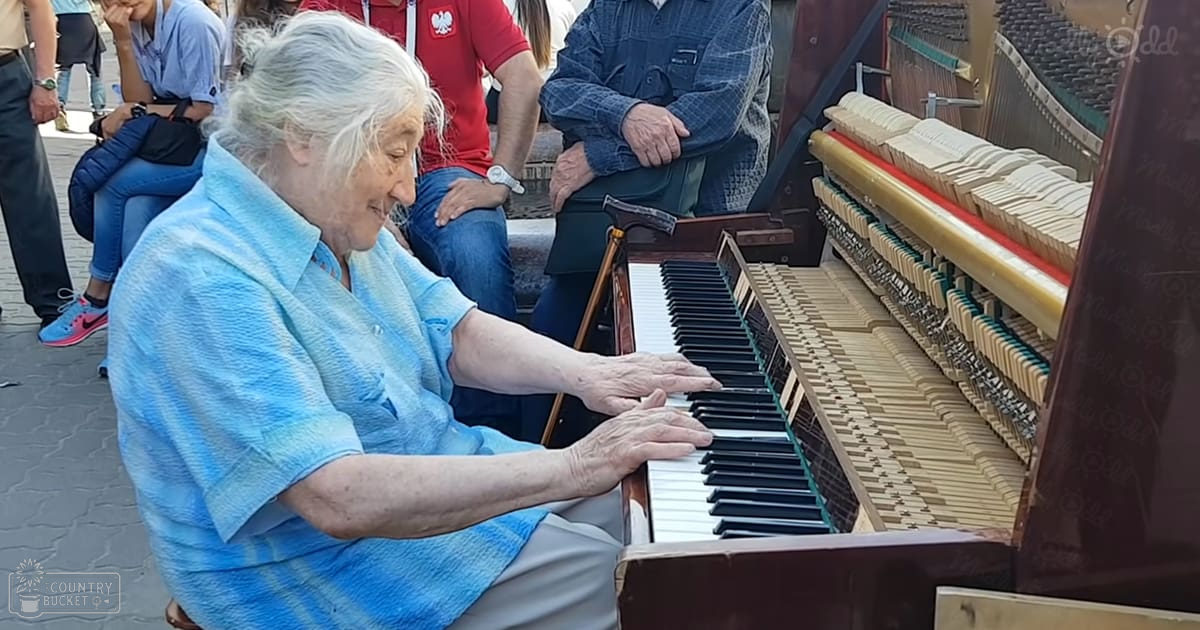 An older adult female playing a grand piano in an outdoor public setting with other people and benches visible.