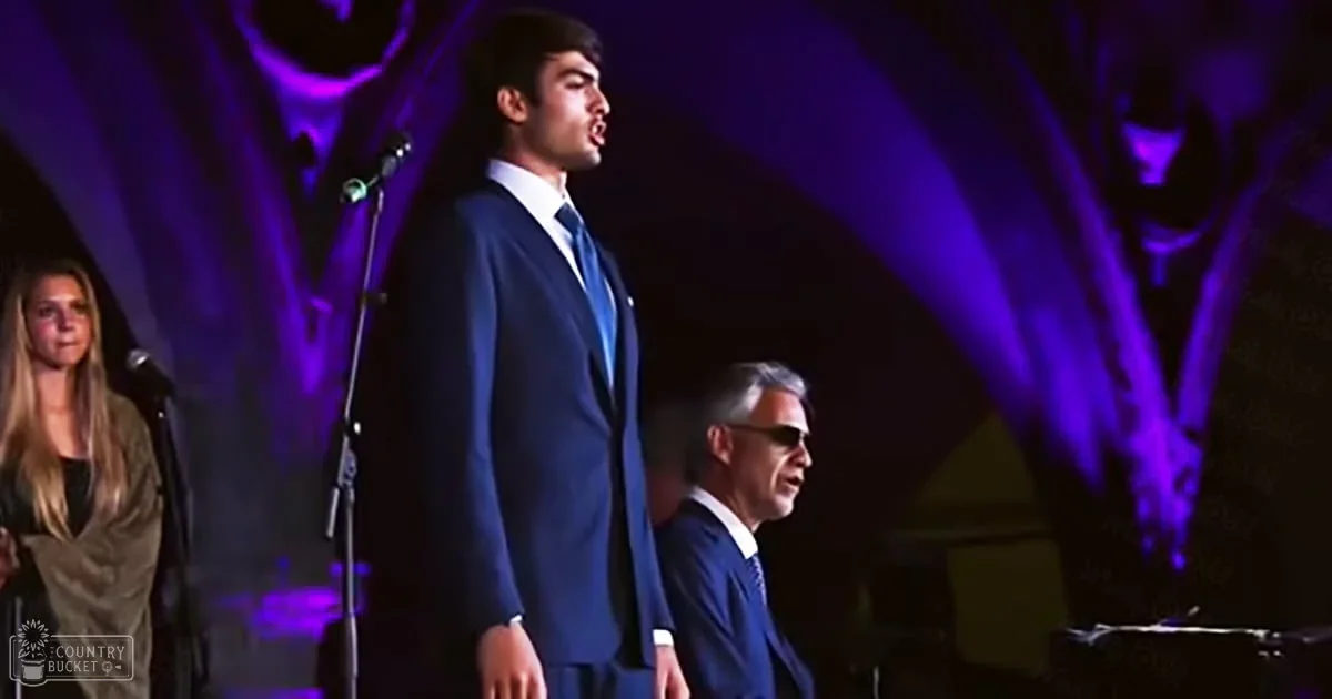A young adult male in a dark blue suit jacket and white dress shirt singing into a microphone at an indoor concert or performance. An adult female in a light brown top with long sleeves is partially visible, while another middle-aged man wearing sunglasses on his head looks on.