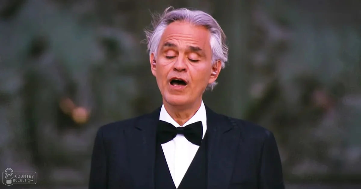 A middle-aged adult male wearing a formal black tuxedo and white shirt, standing outdoors with green foliage in the background.