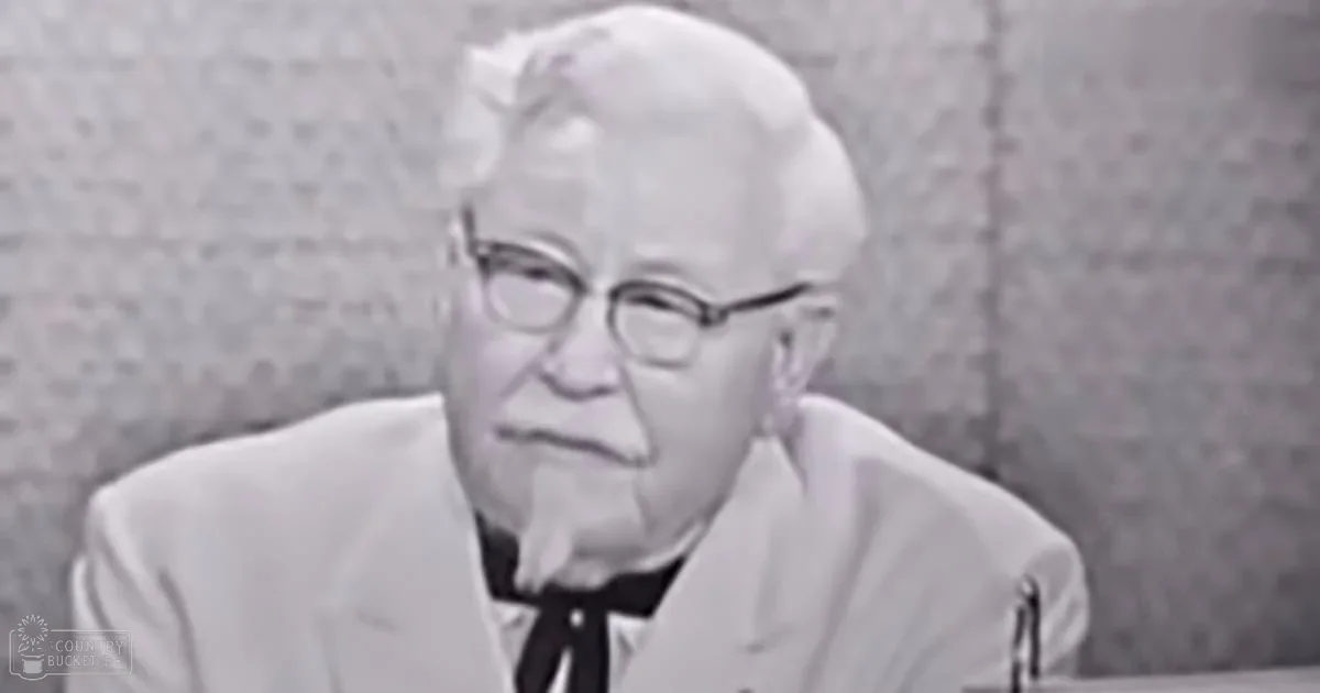 An older adult male in a professional indoor setting, wearing a white suit jacket with black lapels and a black bow tie under a collarless shirt, accessorized with thick-framed glasses and a pen in his ear. The background suggests an office or studio environment.
