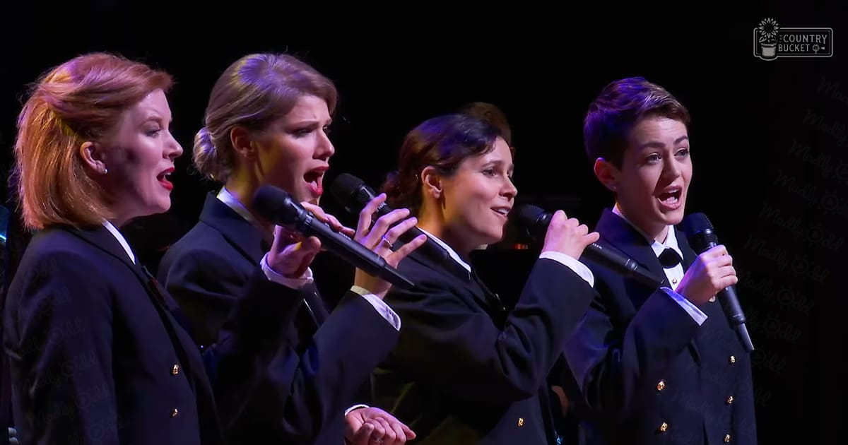 A group of adult female performers singing on stage in a concert setting, wearing dark blazers with gold buttons and white shirts under them.