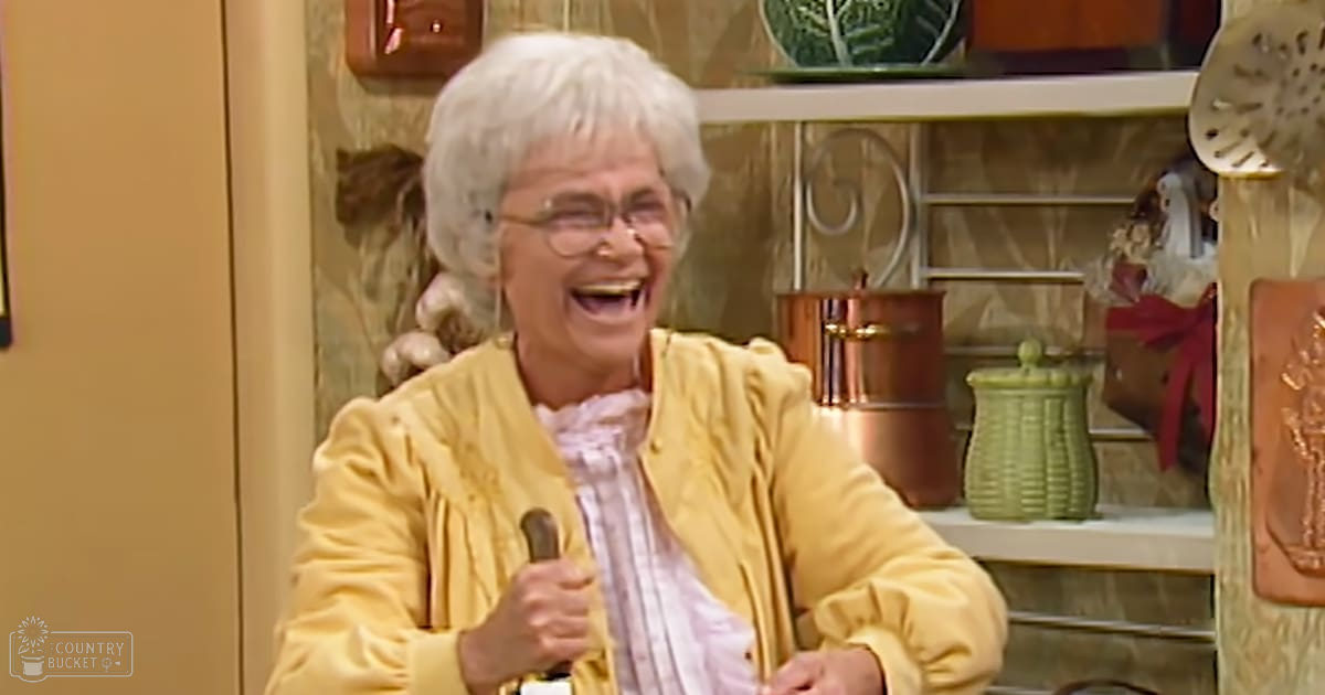 An older adult female wearing a yellow cardigan sweater and collared shirt in a well-decorated kitchen setting with warm lighting.