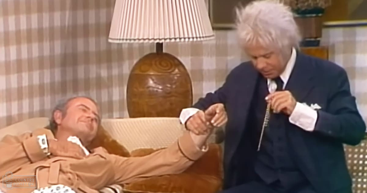 A comedic scene set in an indoor living room with two middle-aged men wearing formal attire; a beige trench coat, dark blue suit jacket, and plaid wallpaper with natural light coming from outside the frame.