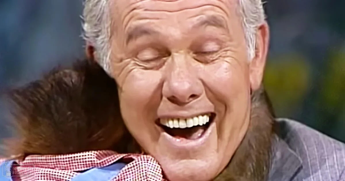 An older adult male wearing a plaid collared shirt, seemingly indoors with muted colors and possibly artificial lighting.