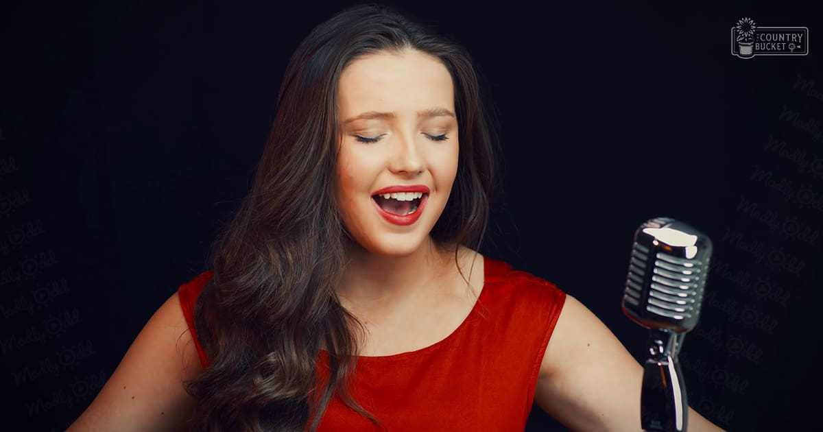 A young adult female in a sleeveless red dress sings into a vintage-style microphone against a dark indoor backdrop, suggesting a recording or performance setting.
