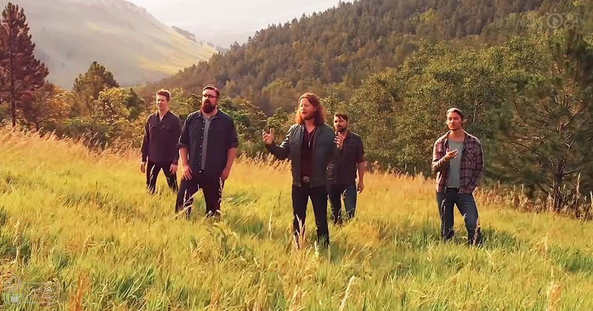 Five young adult males standing in an open grassy field surrounded by dense trees and mountains under soft, natural light at what appears to be either early morning or late afternoon.