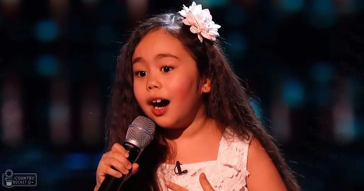 A young child with long dark hair adorned with a white flower accessory sings into a microphone on an indoor stage, illuminated by bright lighting against dimmer background suggesting evening or night time.