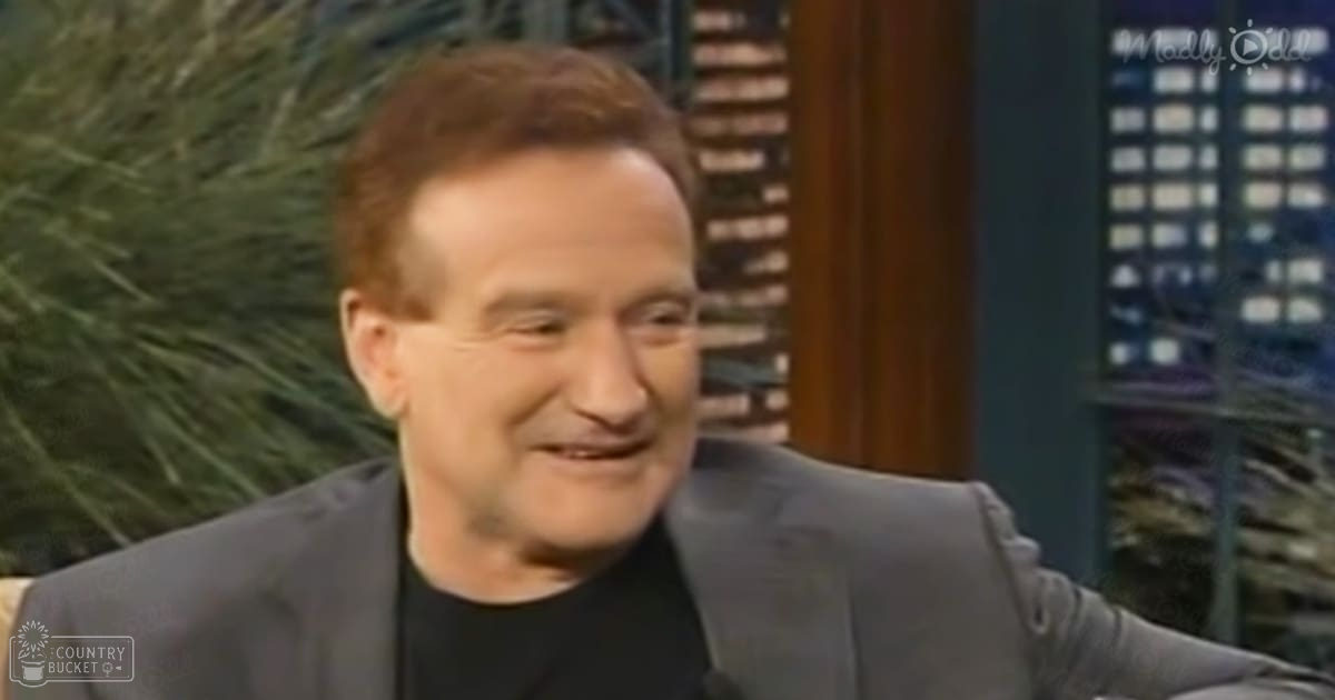 An older adult male wearing a dark gray suit jacket in a studio setting with decorative plants in the background.