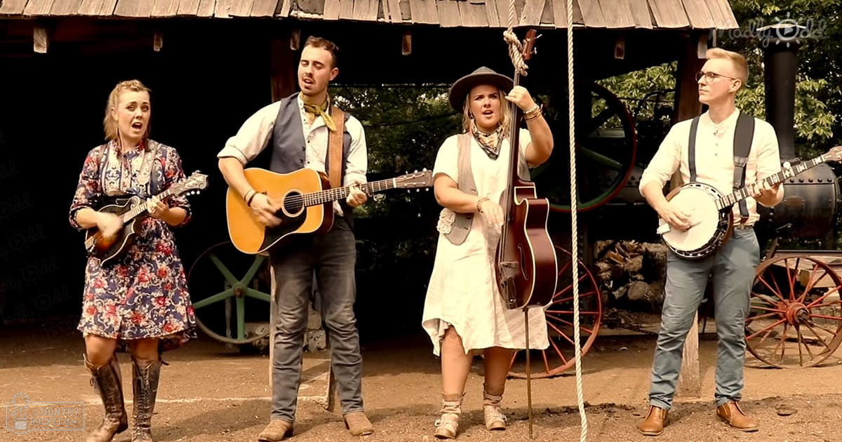 A musical group performing outdoors in a rustic setting with greenery, an old-fashioned steam engine, and earthy tones.