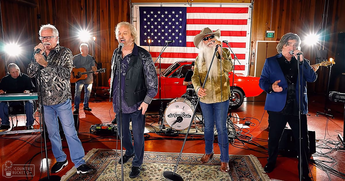 An indoor musical performance with performers wearing casual attire in a setting that includes musical instruments and patriotic decorations.