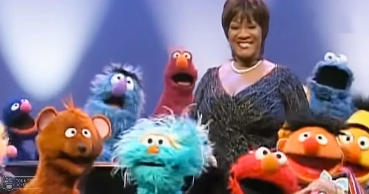 An adult female wearing a dark blue textured dress with a pearl necklace stands among various puppet characters on a stage or performance setting.