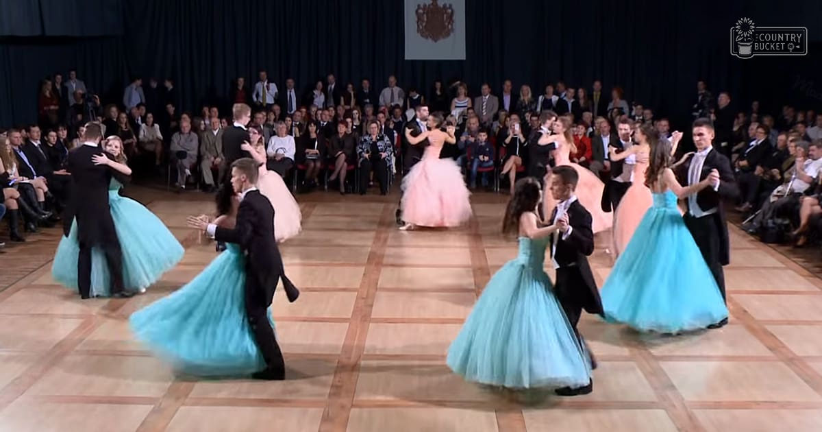 An indoor dance performance with male dancers in black tuxedos and female dancers in flowing turquoise dresses, observed by an audience seated on red chairs under artificial lighting.