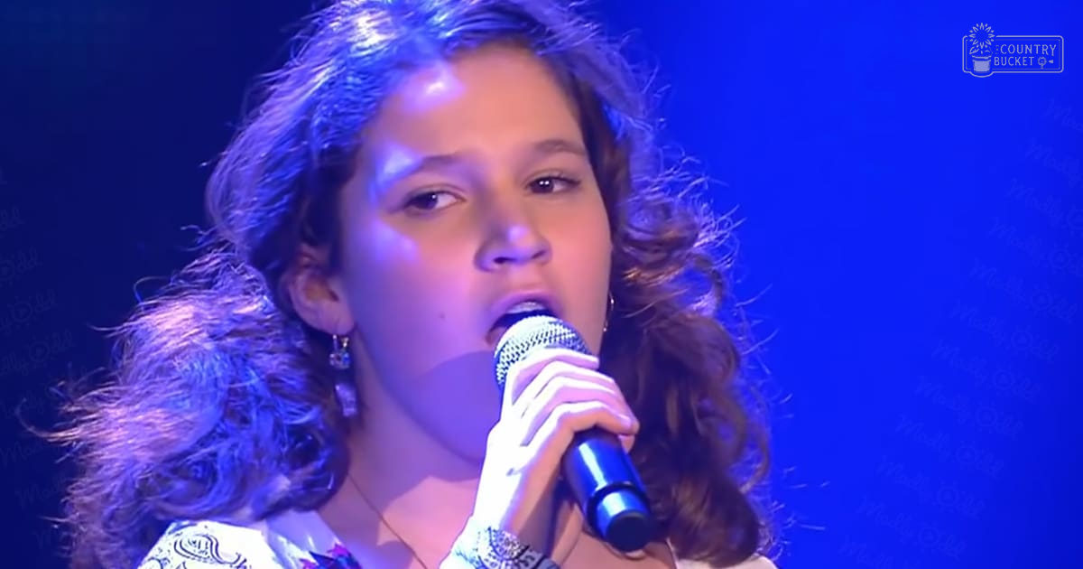 Young adult female singer performing, wearing patterned top with intricate designs in a blue-lit indoor setting holding microphone.