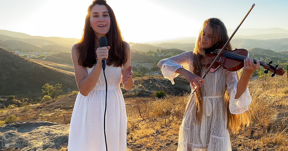 A woman singing outdoors with a scenic background of hills and mountains, accompanied by a child playing the violin under warm late afternoon light.