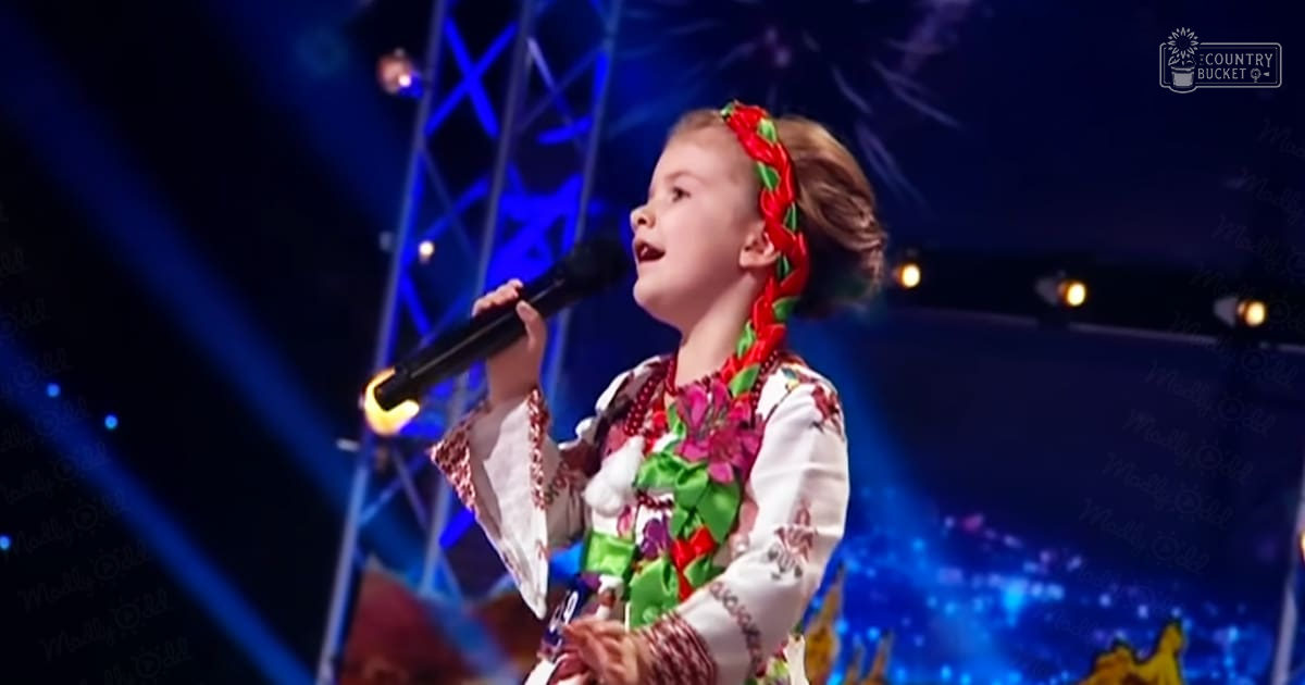 A child, presumably between 5-8 years old and female, performs on stage at an event wearing a traditional-style garment with floral and geometric patterns in bright colors like red, green, yellow, and pink. The setting appears to be indoors on a stage at what seems to be a talent show or similar event.