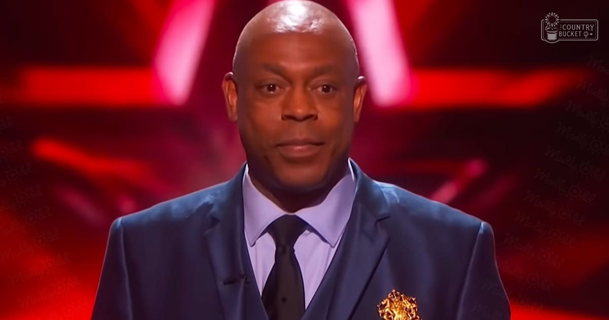 An adult male performer in a dark blue tailored suit jacket with a lapel pin at an indoor performance venue with red lighting.