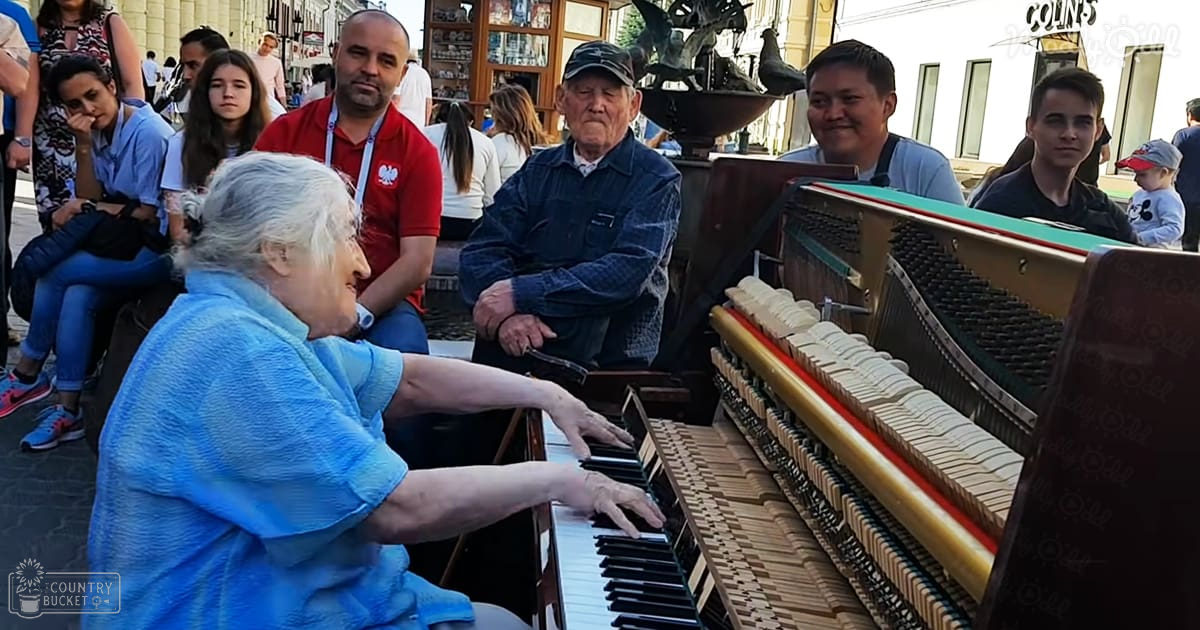 An older adult woman playing a piano with a man in a red polo shirt next to her, surrounded by other adults in a public square during spring or summer with cobblestone pavement and shops.