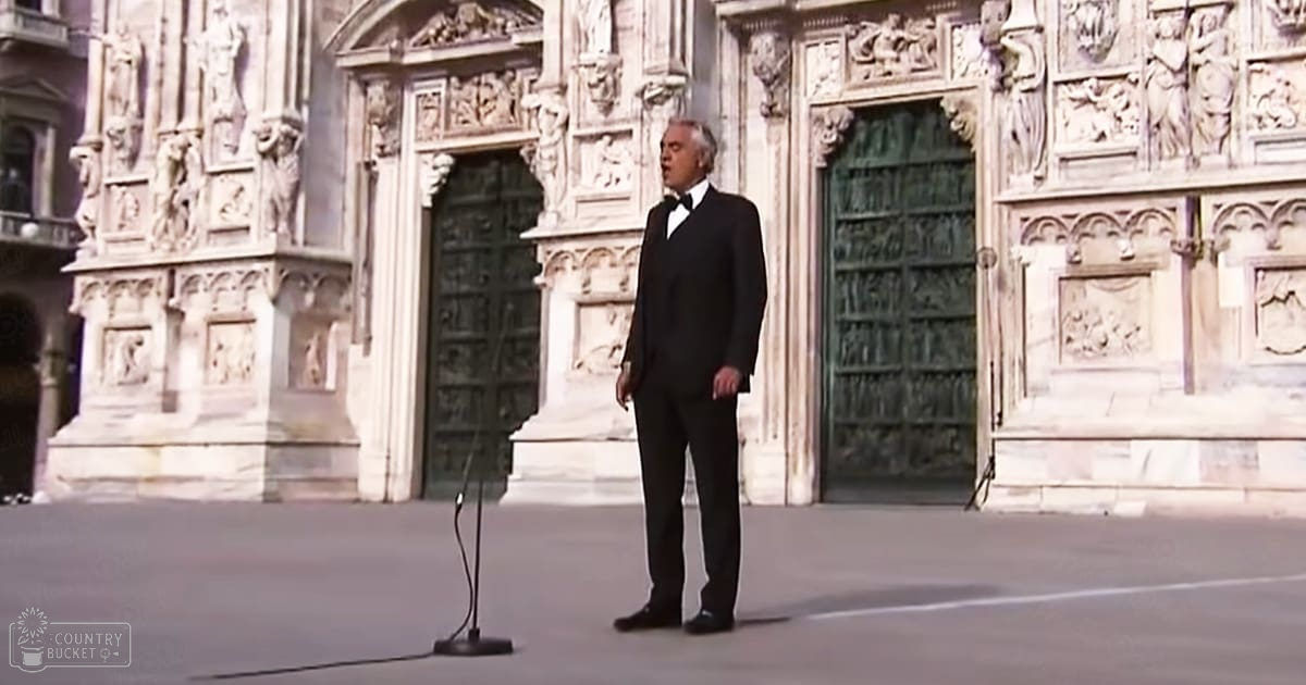 Middle-aged adult male in a formal tuxedo with bow tie, singing at a microphone stand in front of an ornate cathedral-like building under natural daylight.
