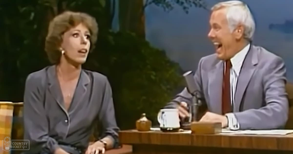 Adult female talk show host wearing a dark gray blazer with deep V-neckline, seated next to an older male guest in a light purple suit jacket and maroon tie. The setting appears to be a studio designed as an outdoor scene.