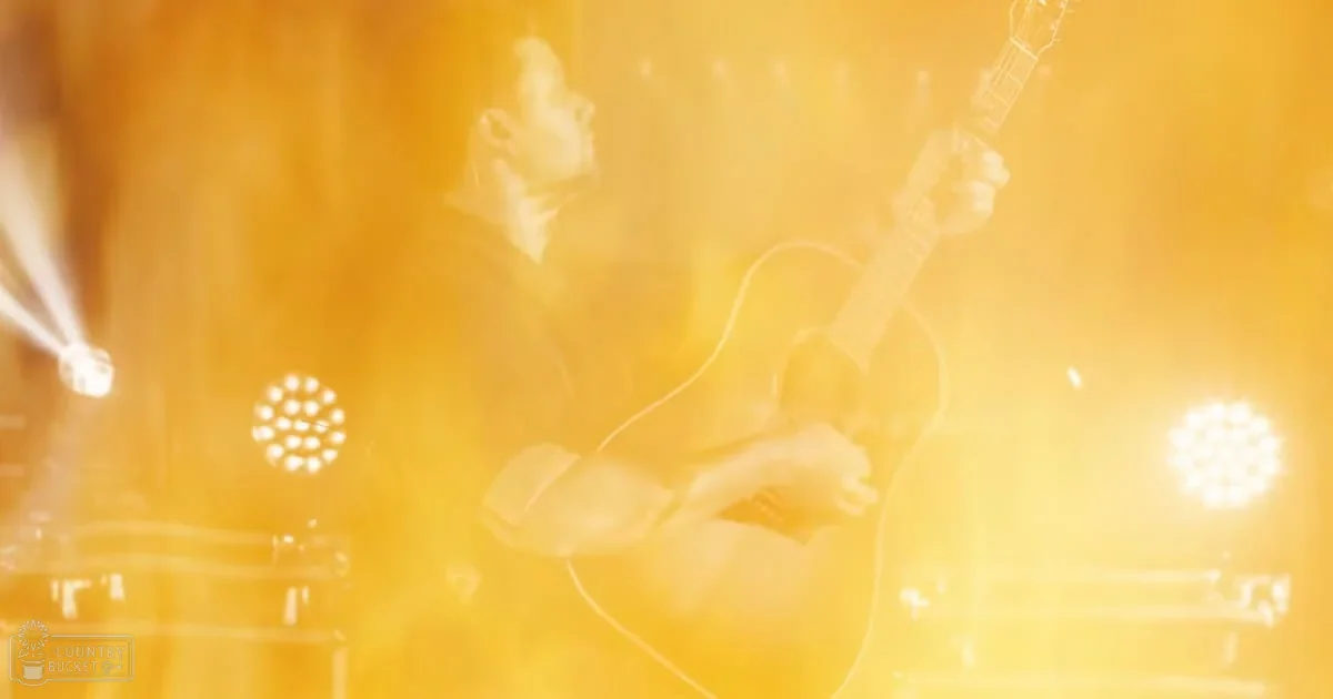 Adult male musician playing an acoustic guitar on stage under warm yellow lights during a concert.