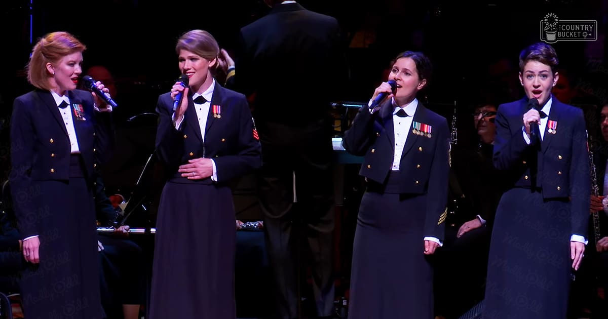 A group of four adult females dressed in dark navy blue military-style uniforms with medals, performing at an event or concert setting.