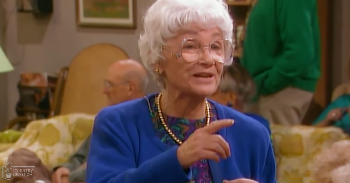An older adult woman wearing a blue cardigan over a patterned blouse with green and purple tones, gold necklace, glasses, in a living room setting with other people present.