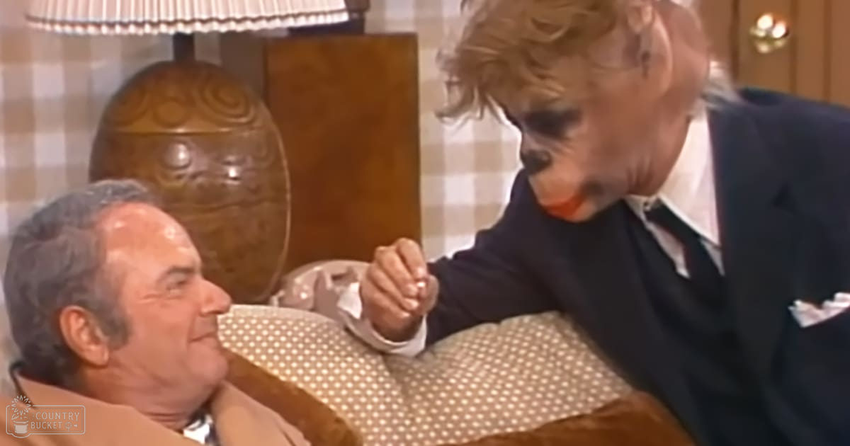 Middle-aged man wearing a light brown blazer and white shirt reclining on furniture with another middle-aged to older adult male in a dark blue suit jacket, plaid wallpaper suggesting mid-20th-century decor, indoors in what looks like a bedroom or living room.
