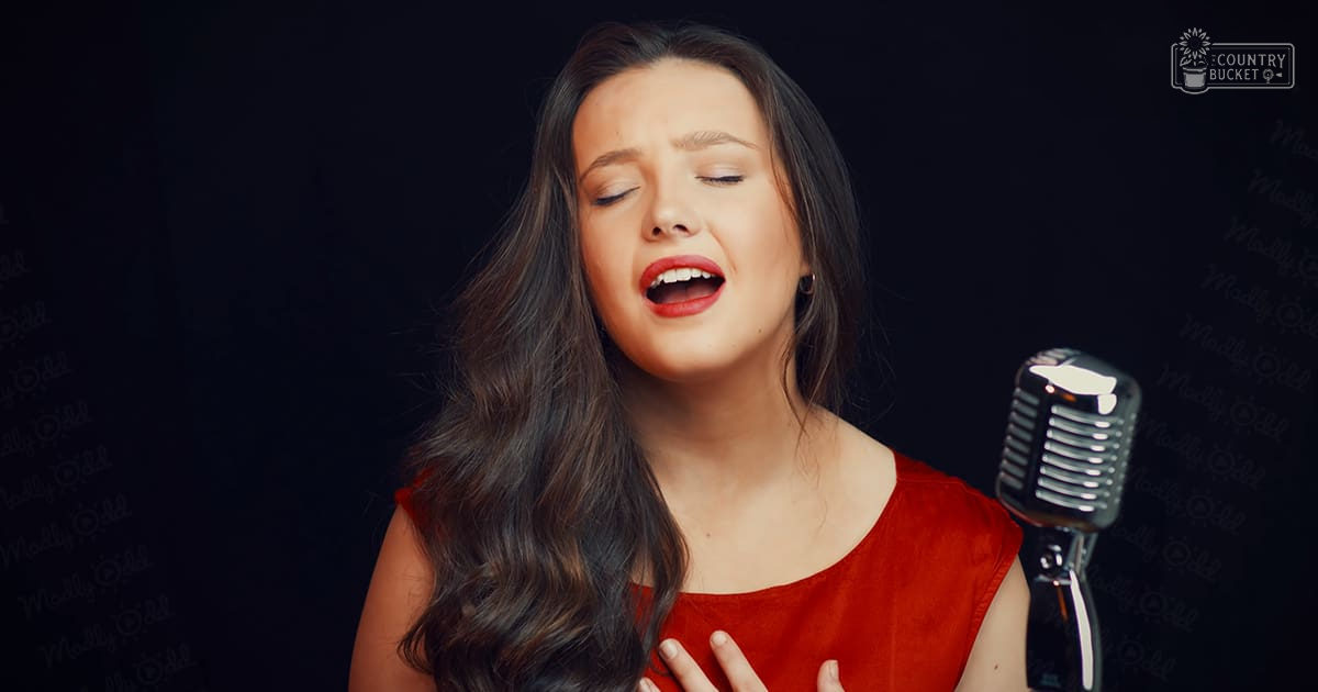 A young adult female singer wearing a vibrant red sleeveless top, holding a vintage-style microphone against an indoor dark backdrop under dramatic lighting.