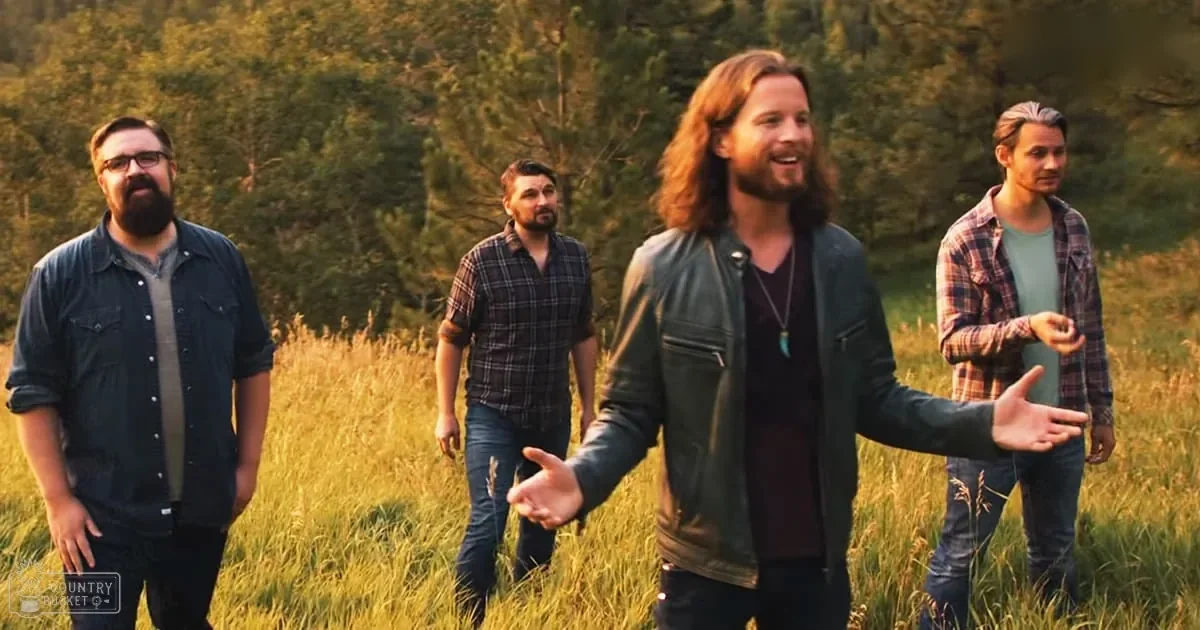 Four adult men in casual attire stand together outdoors on a grassy field with trees in the background during late afternoon light.