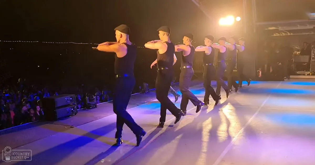 Young adult male dancers performing at a concert hall or theater, wearing black sleeveless tank tops and fitted pants under bright stage lighting with an audience blurred on the left.