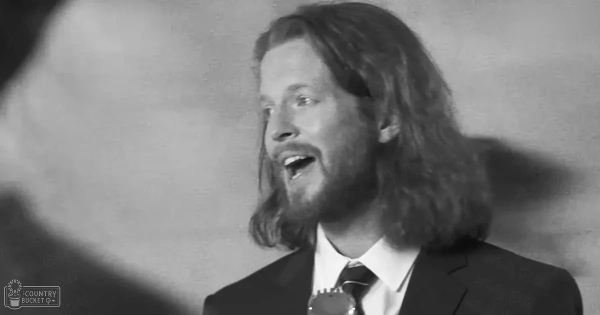 A middle-aged man singing into a microphone indoors with soft lighting; another adult male partially visible in light-colored clothing.