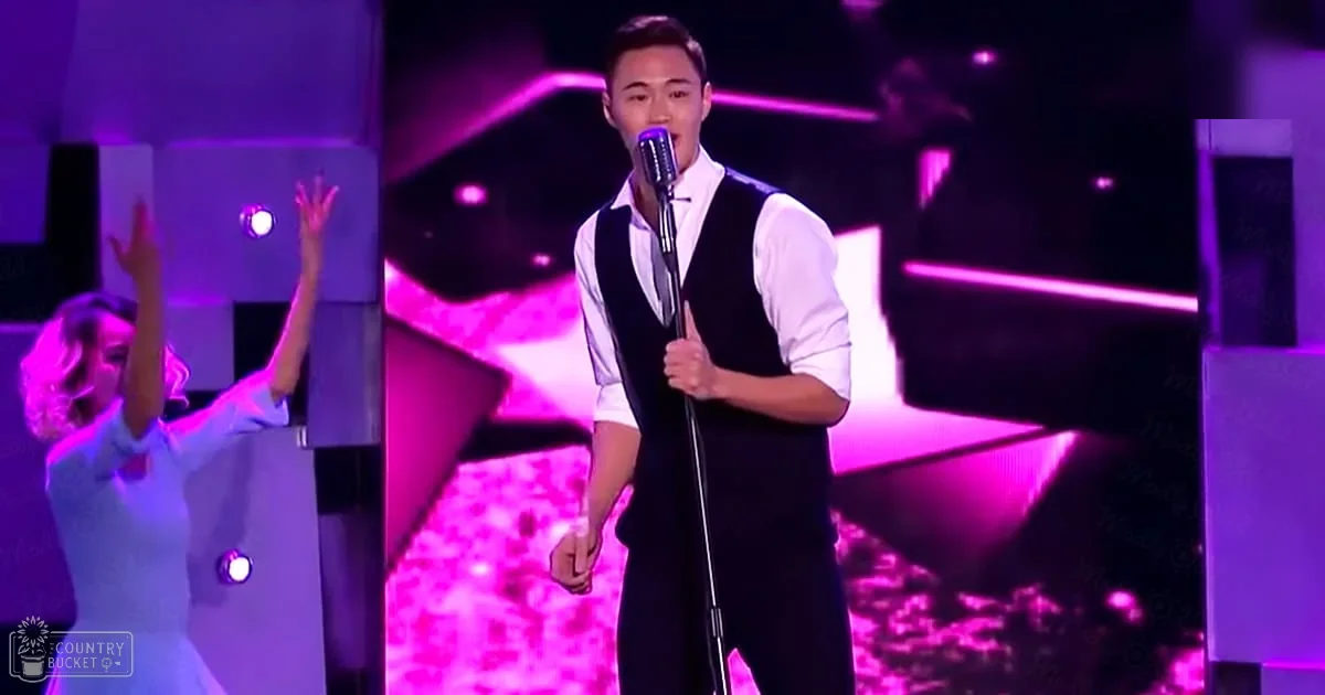 A young adult male singer in a white shirt with rolled-up sleeves and a black vest, performing on stage under vibrant pink and purple lighting.