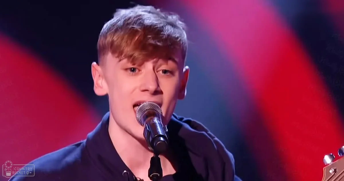 A young adult male singer performing on stage under red lighting, wearing a dark-colored fitted jacket with a high collar.