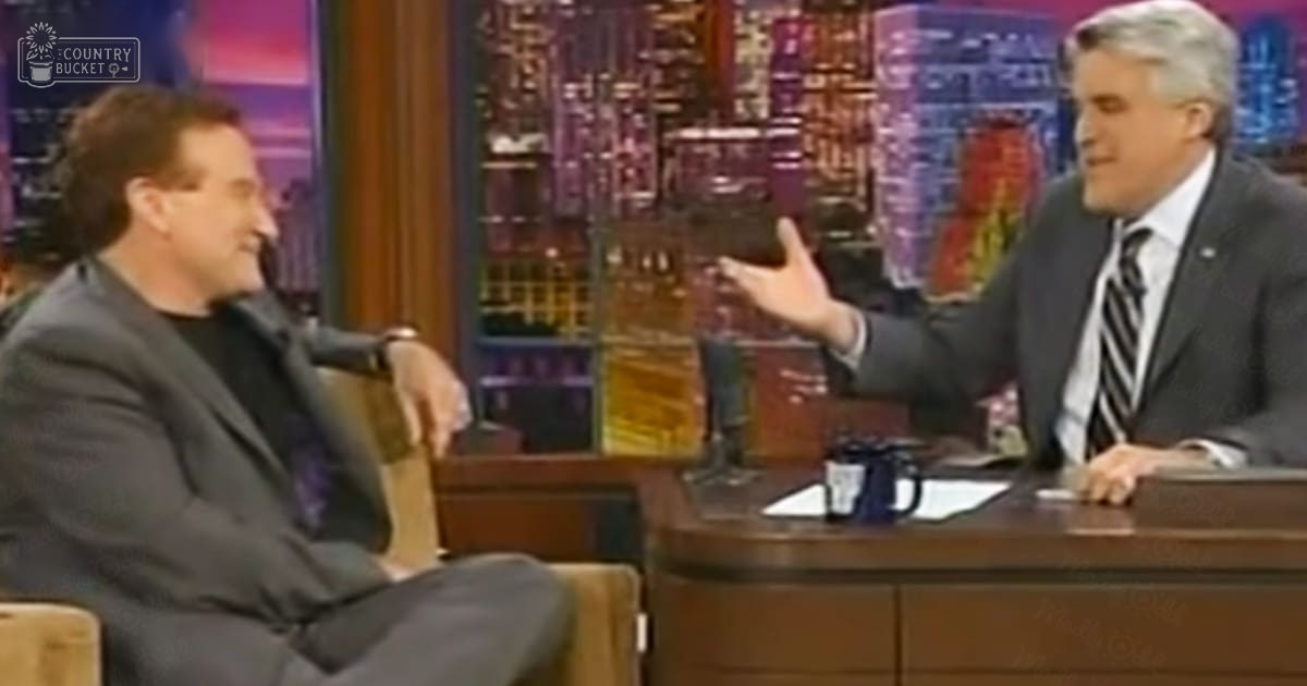 Talk show host and guest seated in a studio with a cityscape backdrop at night, wearing dark suits, illuminated by indoor lighting.