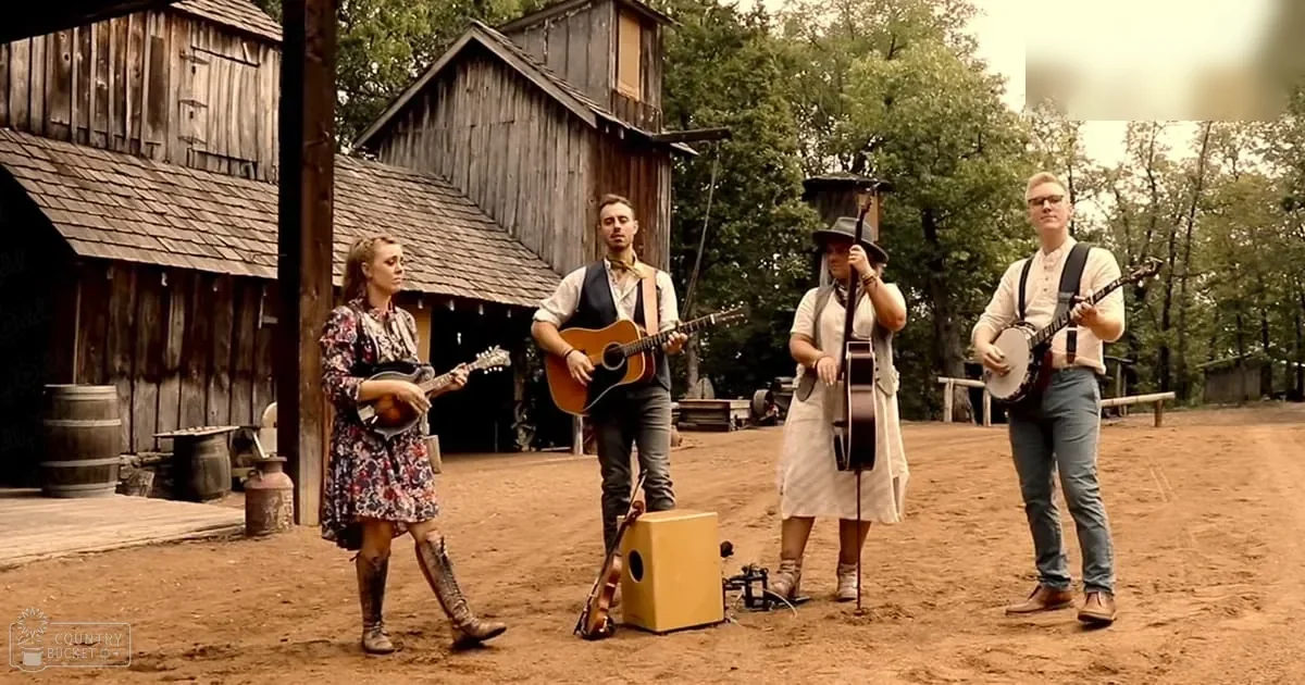 A musical performance set against a frontier town backdrop with young adults dressed in vintage attire playing instruments under natural daylight.