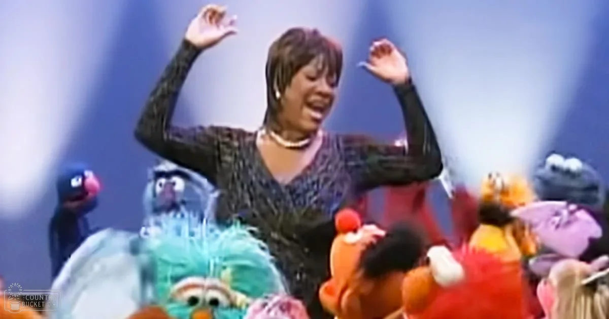 A woman dancing with Sesame Street characters in a studio setting, wearing a black sequined top and pearl necklaces.