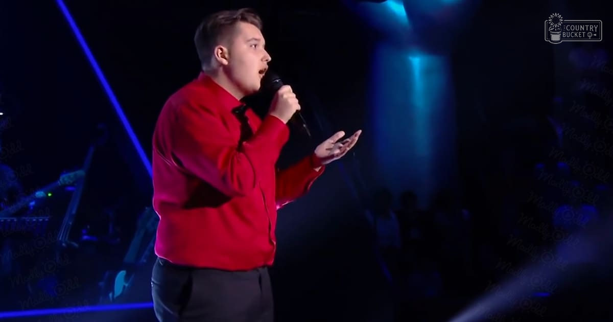A young adult male performing on stage in a bright red long-sleeved shirt with black pants, surrounded by dimly lit blue spotlights at an indoor venue.
