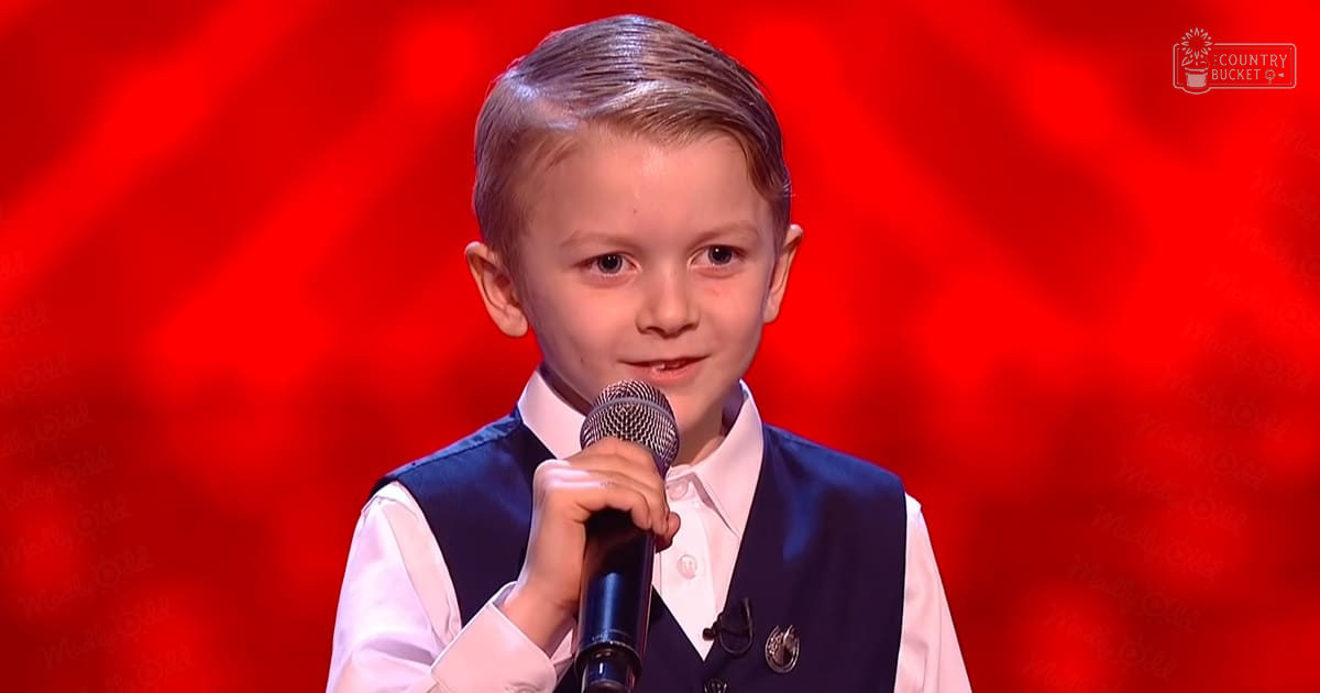 A young child wearing a white dress shirt with a dark blue vest, performing on stage under artificial lighting in an indoor environment.