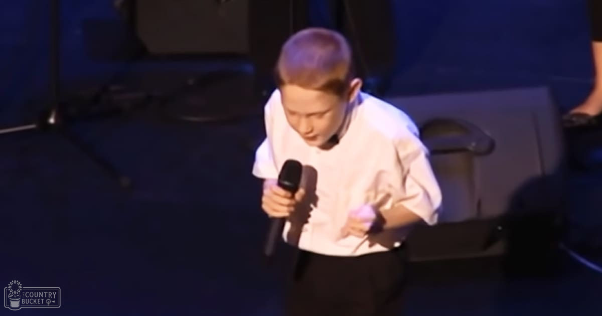 A young child with short hair, wearing a white shirt and dark pants, performing on stage under blue lighting in an indoor venue.