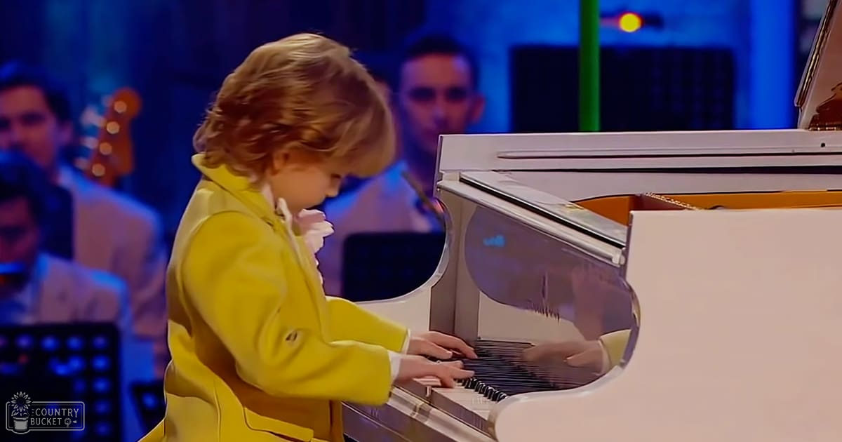 A toddler girl wearing a bright yellow coat with white trim and buttons, playing piano in an indoor venue with stage lighting focused on her.