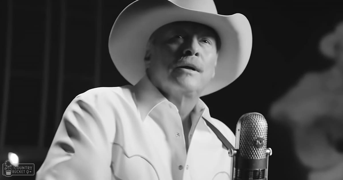 An adult male wearing a classic Western-style white shirt and cowboy hat stands indoors, illuminated by artificial lighting.