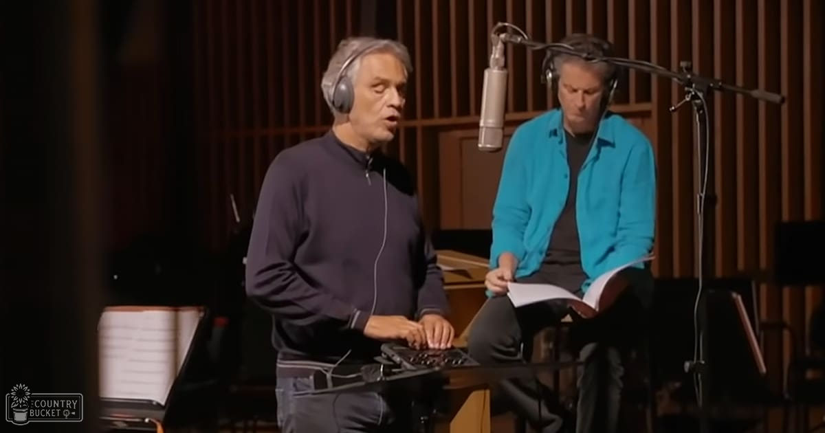 Middle-aged male recording studio session involving music production; middle-aged male wearing dark gray hoodie and headphones, standing near control surface with sheet music stands and soundproofing panels surrounding the setup.