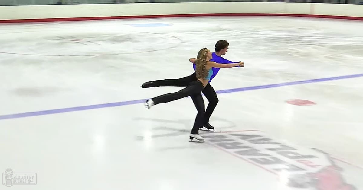 A young adult female figure skater in a blue and black outfit with white ice skates, accompanied by an equally young male figure skater wearing a blue shirt and black pants on an indoor ice rink.