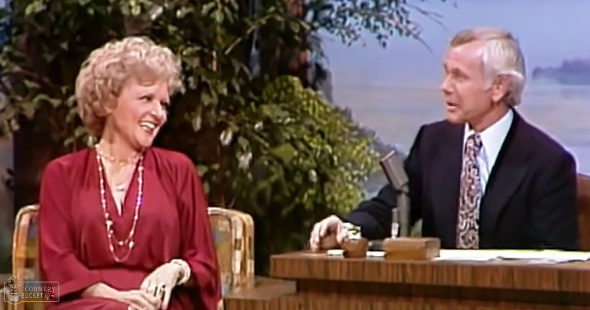 A talk show host in a red blouse and layered pearl necklaces sits with an older male guest wearing a dark suit jacket on a set with greenery in the background, suggesting a daytime television studio.
