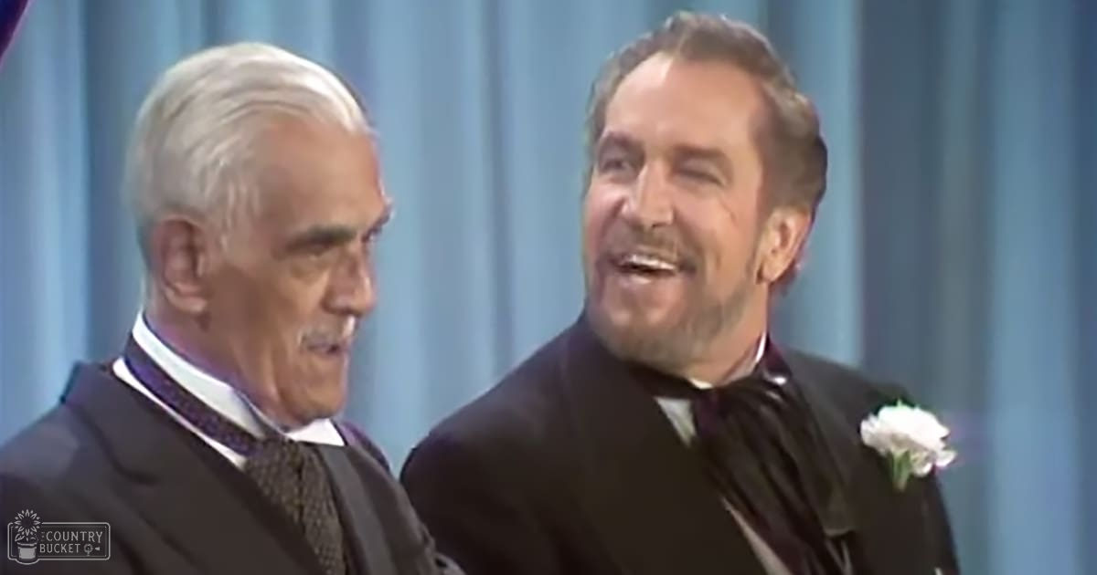 A formal event scene with two older adult males in dark suits, a blue curtain backdrop suggesting an indoor venue.