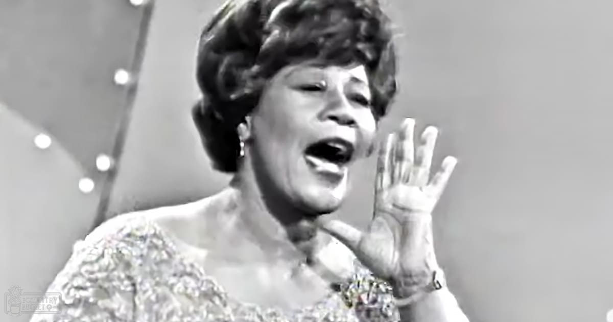 Middle-aged female singing on a stage with decorative lighting, wearing an intricate, glittery mid-20th century-style dress adorned with sequins and embellishments.