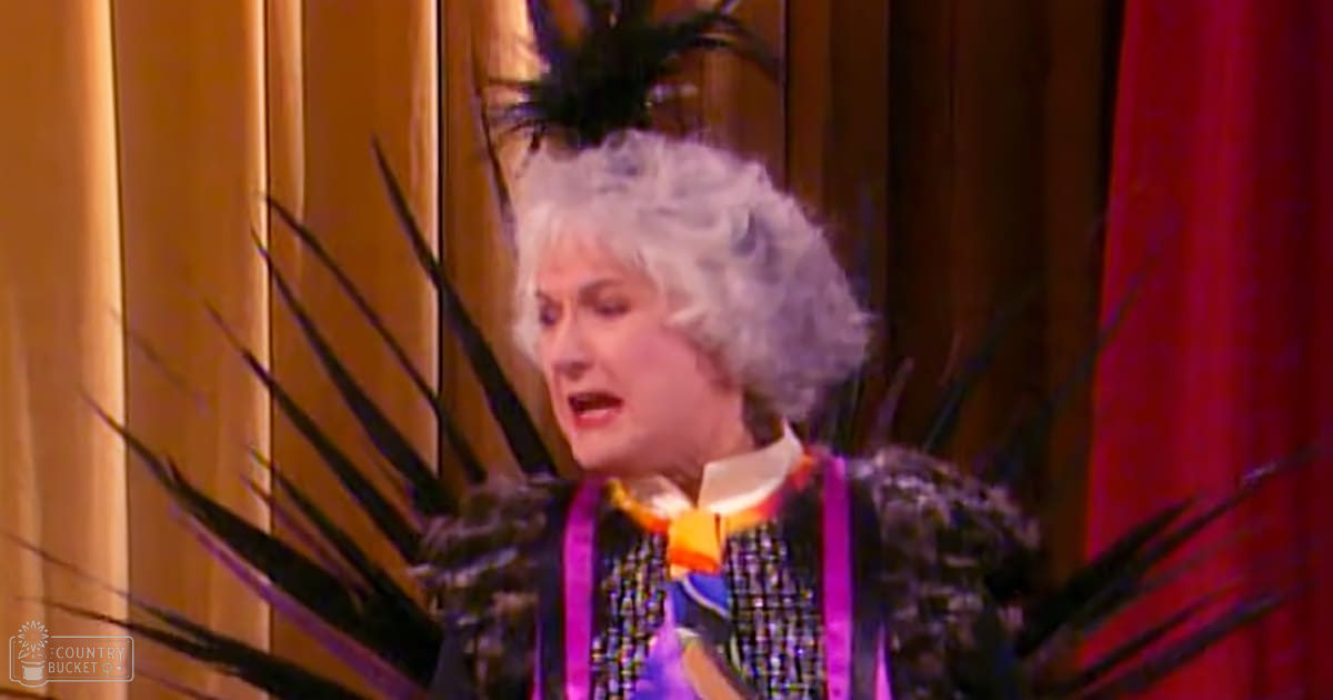 A middle-aged adult female performer in a colorful, patterned outfit with a high collar and feathered headpiece on an indoor stage with wooden paneling and red curtains.